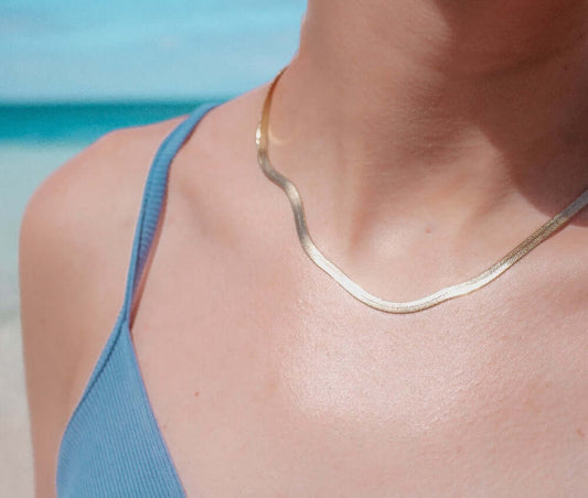 Sand + Salt's herringbone necklace shown on a woman's neck, she is wearing a blue dress standing by Caribbean water. Gold-Filled and Gemstone jewelry, Sand and Salt Studio