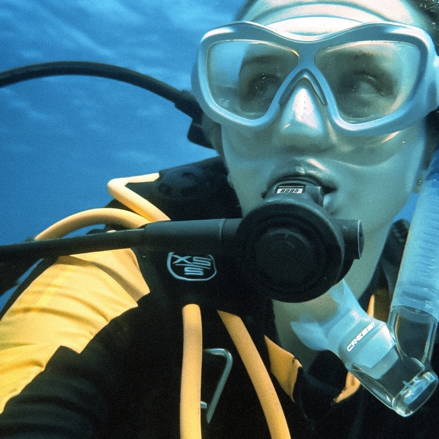 An underwater close-up of a scuba diver wearing a mask, regulator, and yellow-and-black diving suit against clear blue water.