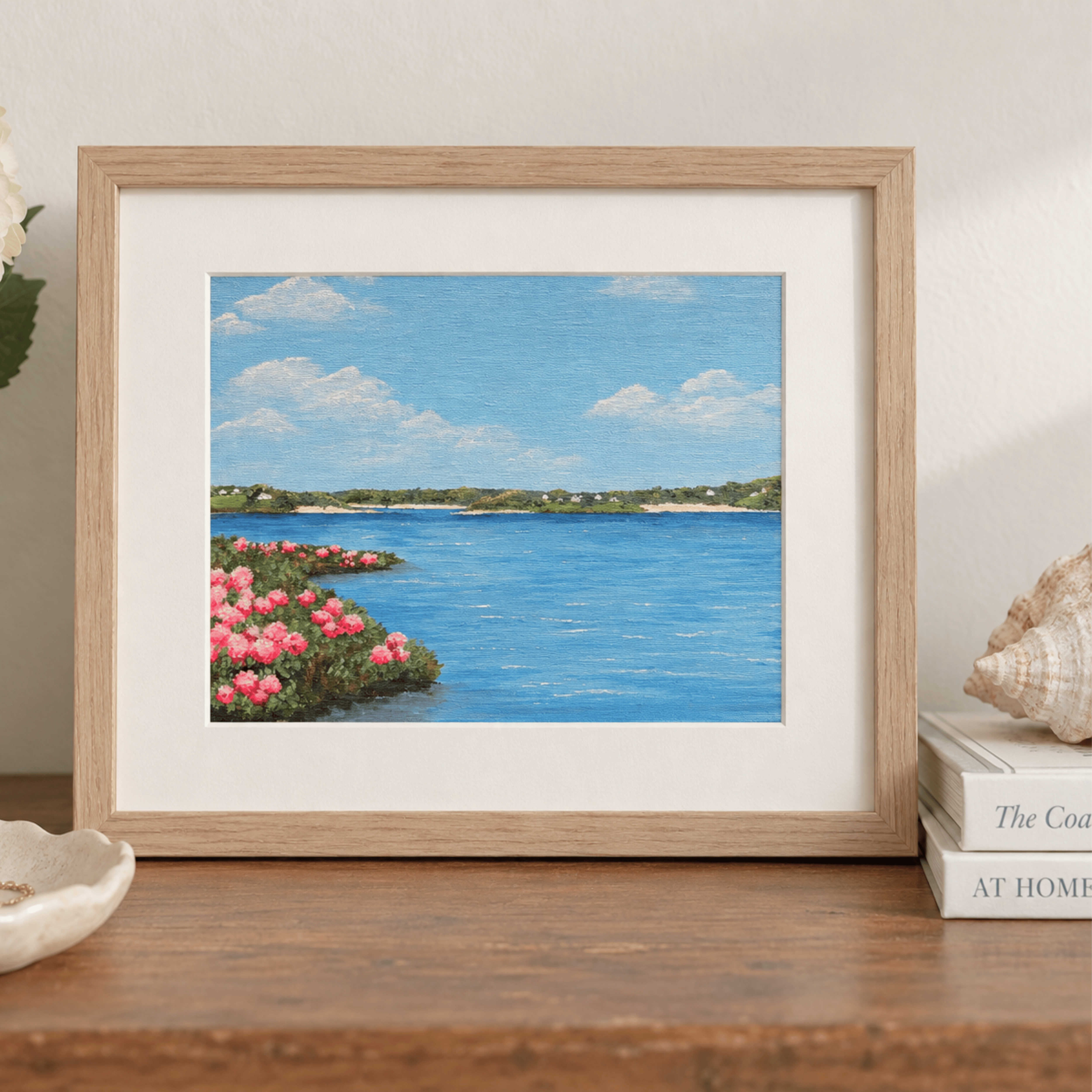 A framed landscape art print depicting a coastal scene with blue water, pink flowers, and a green shoreline under a blue sky, displayed on a wooden shelf next to books and a seashell.