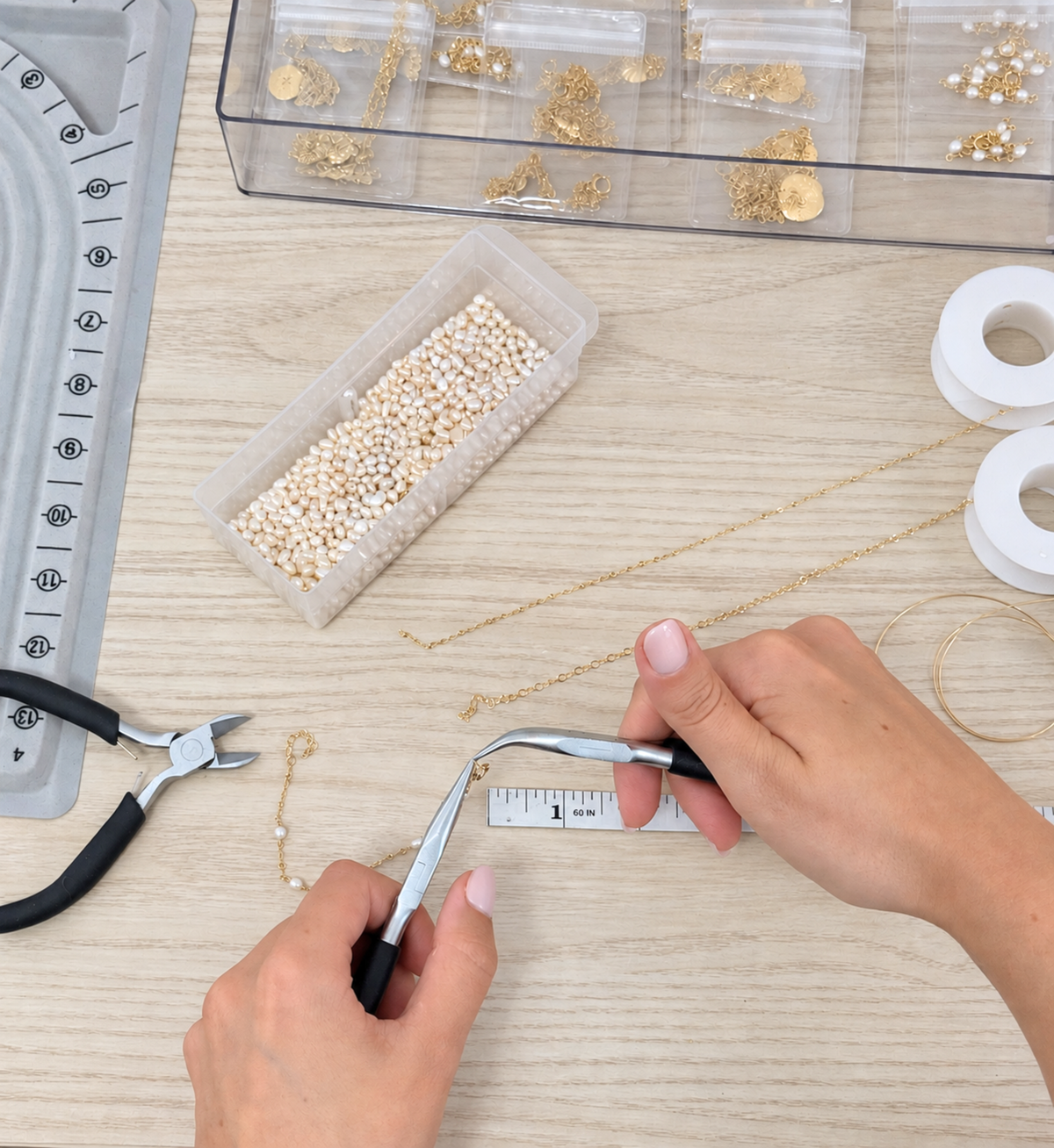 overhead view of hands holding two jewelry pliers and finishing a bracelet, showing pearls, gold chain, and other jewelry making materials